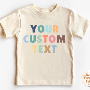 以下が含まれることがあります： クリーム色の半袖Tシャツ。カラフルなレトロスタイルのブロックレターで「YOUR CUSTOM TEXT」と書かれています。丸いネックラインと、右下に小さな円形のロゴが付いています。