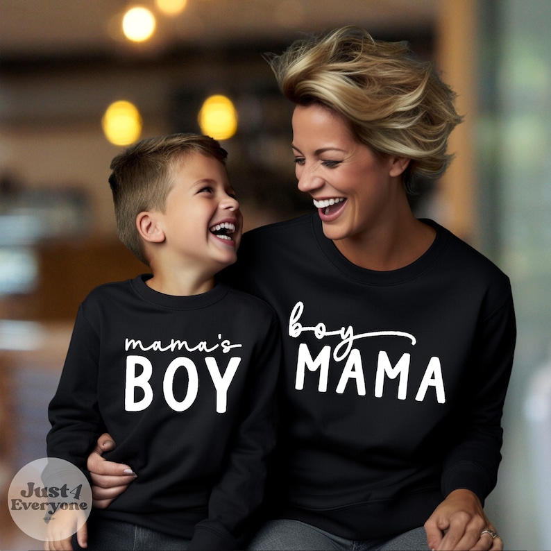May include: Black sweatshirts with white text. One says "mama's BOY" and the other says "boy MAMA". The sweatshirts are worn by a mother and son who are smiling at each other. The image is taken indoors.