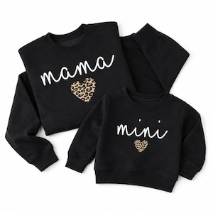 Puede incluir: Dos sudaderas negras de manga larga con cuello redondo. La sudadera más grande presenta el texto blanco "mama" sobre un corazón con estampado de leopardo. La más pequeña presenta el texto blanco "mini" sobre un corazón de leopardo más pequeño.