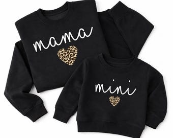 Può includere: Due felpe nere a maniche lunghe con girocollo. La felpa più grande presenta la scritta bianca "mama" sopra un cuore con stampa leopardata. La felpa più piccola presenta la scritta bianca "mini" sopra un cuore leopardato più piccolo.