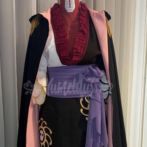 Può includere: Un costume esposto su un manichino composto da un mantello nero con fodera rosa chiaro e dettagli dorati sulle spalle. L'outfit include un top bianco con colletto a balze rosso scuro, una gonna nera con motivi di teschi solari e una fascia color lavanda.