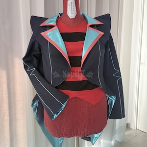 Pode incluir: Um casaco azul-marinho escuro com lapelas e acabamentos em cetim azul-claro, usado sobre uma camisa de riscas horizontais vermelhas e pretas e uma saia vermelha texturizada. O casaco tem ombros estruturados e bainha assimétrica. O texto Yumetly é visível.