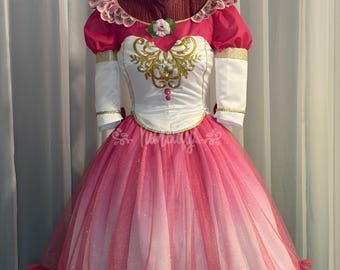 May include: A pink and white dress featuring a white bodice with gold embroidery, short puffed sleeves, and a layered tulle skirt. The dress includes a pink ruffled neckline, a central flower accent, and white elbow-length sleeves with gold trim.