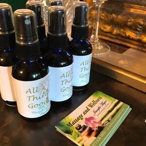 May include: Five small blue glass bottles with black caps and white labels that read "All Things Good Bug and Mosquito Repellent". The bottles are sitting on a brown surface next to a business card that reads "Massage and Wellness Jennifer Major Licensed Massage Therapist 973 828-5852 jerseybeecrazy@gmail.com".