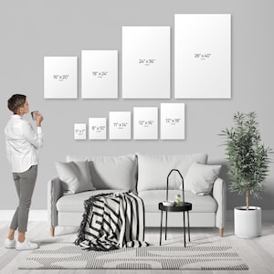 A woman is standing in a living room, looking at a wall with a series of white canvas prints of varying sizes, each labeled with its corresponding dimensions.