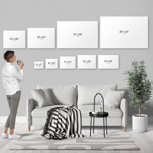 A person standing in a living room, looking at a wall with multiple blank canvas prints of different sizes, and a couch, a coffee table, and a potted plant in the background.