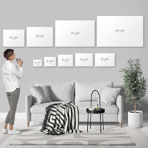 A person standing in a living room, looking at a wall with multiple blank canvas prints of different sizes, and a couch, a coffee table, and a potted plant in the background.