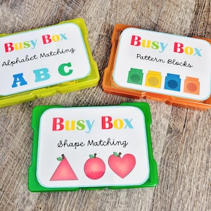 May include: Three colorful Busy Box educational sets. The sets are in green, orange, and yellow containers. Each set has a white label with the words "Busy Box" and a different learning activity: Alphabet Matching, Pattern Blocks, and Shape Matching.
