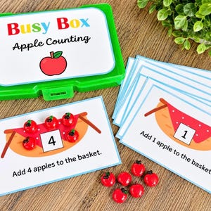 May include: A green plastic storage box labeled Busy Box Apple Counting with a stack of white activity cards featuring illustrations of baskets and apples. The cards include text instructions and numbers, accompanied by small red apple-shaped tokens.