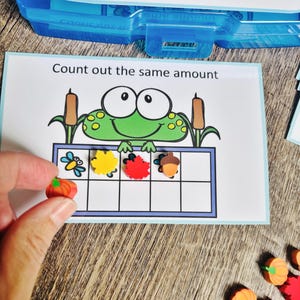 May include: A rectangular educational card with the text Count out the same amount, featuring a green frog illustration above a ten-frame grid. The grid contains small colorful erasers shaped like a blue dragonfly, a yellow leaf, a red leaf, and an acorn.