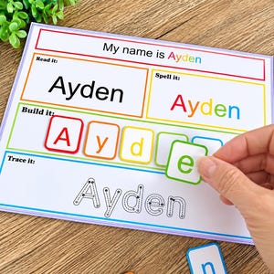May include: A rectangular educational worksheet titled My name is Ayden with sections for reading, spelling, building, and tracing the name. The build section features colorful square letter tiles, with a hand placing a green tile with the letter e.