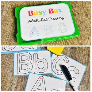 Puede incluir: Una caja de pl&aacute;stico verde con la etiqueta "Busy Box Alphabet Tracing" contiene tarjetas para trazar el alfabeto. Las tarjetas presentan l&iacute;neas punteadas para trazar las letras may&uacute;sculas y min&uacute;sculas A, B y C. Tambi&eacute;n se ve un rotulador negro.