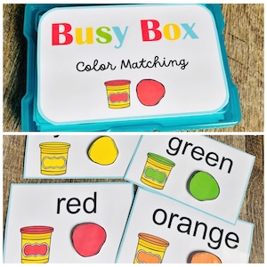 Puede incluir: Una caja de pl&aacute;stico azul con la etiqueta "Busy Box Color Matching" contiene tarjetas de combinaci&oacute;n de colores. Las tarjetas presentan ilustraciones de botes de plastilina y bolas de plastilina de colores a juego. Las tarjetas individuales muestran colores como rojo, verde y naranja.