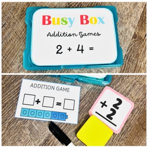 Puede incluir: Un contenedor de pl&aacute;stico azul con la etiqueta "Busy Box Addition Games" con la ecuaci&oacute;n 2 + 4 = mostrada. Tarjetas adicionales muestran un juego de suma con casillas y una tarjeta num&eacute;rica con el n&uacute;mero 2. Tambi&eacute;n se ve un marcador negro y una goma de borrar amarilla.