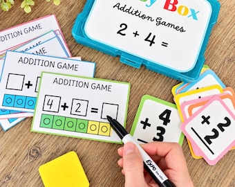 May include: A teal plastic storage case labeled Busy Box Addition Games 2 + 4 = with multiple colorful addition game cards, a yellow square eraser, and a black dry-erase marker used to complete math problems on the cards.
