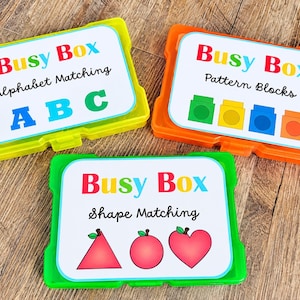 May include: Three rectangular plastic storage containers in lime green, orange, and bright green. Each lid features a white label with the text Busy Box, followed by specific activity titles: Alphabet Matching, Pattern Blocks, and Shape Matching.