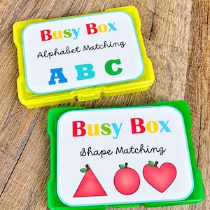 May include: Two rectangular plastic storage containers, one yellow and one green, each featuring a white label. The yellow box is labeled Busy Box Alphabet Matching with blue, green, and red letters. The green box is labeled Busy Box Shape Matching with red shapes.