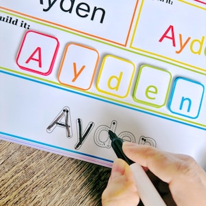 Pu&ograve; includere: Un foglio di lavoro bianco con il nome "Ayden" scritto in nero in alto. Il foglio di lavoro ha una sezione per costruire il nome con tessere di lettere, una sezione per tracciare le lettere e una sezione per scrivere il nome. Le lettere A, y, d, e e n sono scritte in nero sul foglio di lavoro. Una mano tiene un pennarello nero e traccia le lettere.