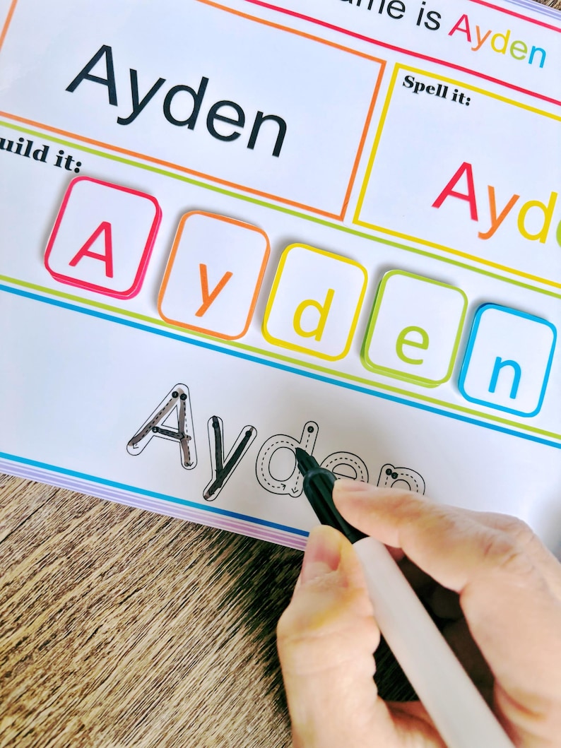 Pu&ograve; includere: Un foglio di lavoro bianco con il nome "Ayden" scritto in nero in alto. Il foglio di lavoro ha una sezione per costruire il nome con tessere di lettere, una sezione per tracciare le lettere e una sezione per scrivere il nome. Le lettere A, y, d, e e n sono scritte in nero sul foglio di lavoro. Una mano tiene un pennarello nero e traccia le lettere.