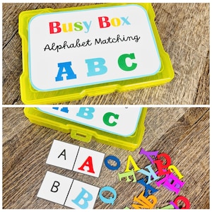 Puede incluir: Una caja Busy Box de pl&aacute;stico amarillo para emparejar el alfabeto. La caja contiene tarjetas con las letras A, B y C, y letras individuales. La tapa de la caja tiene las palabras "Busy Box Alphabet Matching" en rojo, azul y verde.