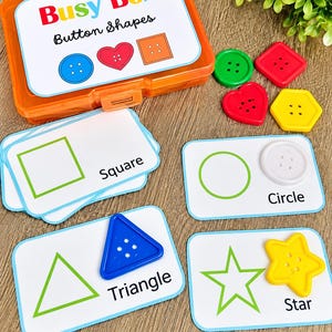 May include: An orange plastic Busy Box labeled Button Shapes containing colorful plastic buttons in circle, heart, square, triangle, star, and hexagon shapes, alongside matching white activity cards with shape outlines and names.