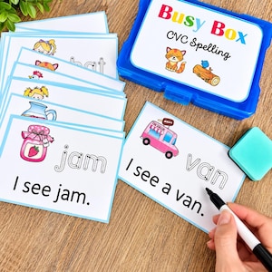 May include: A blue plastic storage box labeled Busy Box CVC Spelling with a stack of white educational cards featuring colorful illustrations and text. The cards show words like jam and van with dotted letters for tracing and sentences like I see jam.