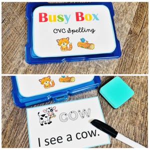 Puede incluir: Un kit azul de pl&aacute;stico Busy Box CVC Spelling con una tarjeta blanca con un gato de dibujos animados y un p&aacute;jaro en un tronco. Una tarjeta muestra la palabra "COW" y la frase "I see a cow". Tambi&eacute;n hay un borrador turquesa y un rotulador negro.