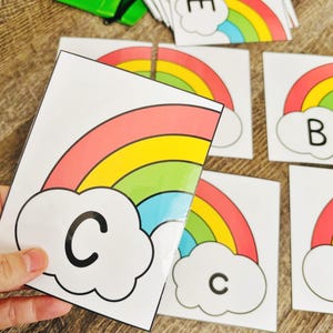 May include: Educational flashcards featuring colorful rainbows and letters. The cards have a white background with a rainbow design in red, orange, yellow, green, and blue. Each card has a letter in a cloud.