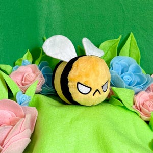 May include: A plush bee toy with a yellow body and black stripes, white wings, and a stern expression. The bee is surrounded by felt flowers in shades of pink and blue, with green leaves, set against a green backdrop.