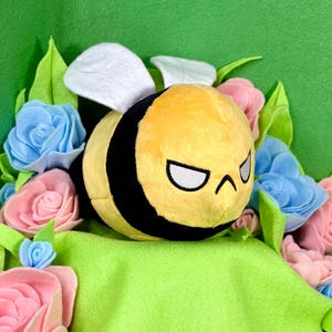 May include: A plush bee toy with a yellow body, black stripes, and white wings, surrounded by felt flowers in shades of pink and blue. The bee has an angry expression. The background is a solid green.