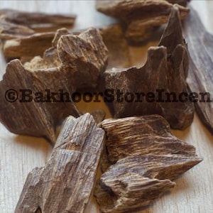 May include: Close-up of several pieces of dark brown oud wood, with a rough, textured surface. The pieces are irregularly shaped and scattered on a light-colored wooden surface. The text "©BakhoorStoreIncense" is visible.
