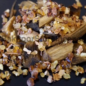 May include: Close-up of a pile of raw incense ingredients. The image features light brown wood chips and amber-colored resin pieces. The ingredients are scattered on a dark surface, creating a textured appearance.