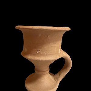 May include: A terracotta chalice with a wide, flared rim, a narrow stem, and a circular base. The vessel features a single curved handle attached to the side, with a matte, light brown finish and visible surface texture throughout the clay body.