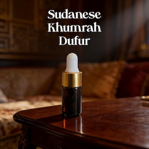 May include: A small dark amber glass bottle with a gold collar and a white dropper top. The text Sudanese Khumrah Dufur is printed in white above the bottle. The bottle is cylindrical with a smooth finish and a rounded white applicator tip.