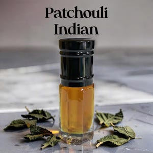 Puede incluir: Un frasco pequeño de vidrio transparente lleno de líquido color ámbar, con una tapa negra. El texto "Patchouli Indian" aparece impreso sobre el frasco. Varias hojas verdes secas y dentadas están dispuestas alrededor de la base del envase.