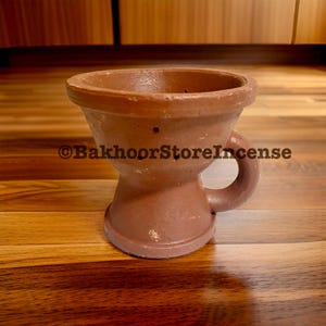 May include: A brown terracotta incense burner with a handle. The burner has a wide top and a rounded base. The text "©BakhoorStoreIncense" is printed on the side. It is placed on a wooden surface. A decorative item for the home.