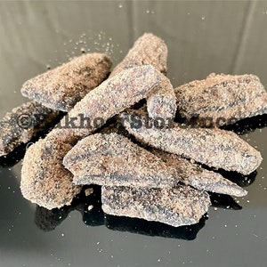 Può includere: Un mucchio di pezzi di frutta secca di colore marrone scuro ricoperti di zucchero di canna chiaro. I pezzi di frutta sono disposti in modo casuale su una superficie nera.