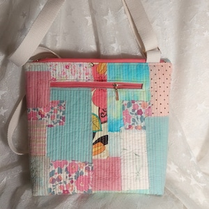 以下が含まれることがあります： 薄い青、ピンク、白の花柄の、パッチワークのクロスボディバッグ。バッグには長い白いストラップと金色のジッパーが付いています。