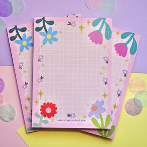 May include: Three pink notepads with a grid pattern and a floral border. The border features flowers in shades of blue, pink, and red. The notepads have a scalloped edge and the text "www.meggarrodart.com" printed on the bottom.
