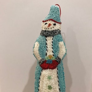 May include: A blue and white snowman ornament with a red hat and a black and white scarf. The snowman is holding a red basket with colorful balls inside.