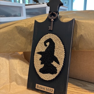 May include: A dark gray wooden plaque with a witch silhouette and the text "Salem 1692". The plaque has a decorative key and a gold-colored oval design. The plaque is approximately 10 inches tall.