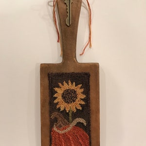 May include: A wooden paddle with a brown, orange, and yellow sunflower and pumpkin design. The paddle has a key with the word "LIFE" on it attached to the top.