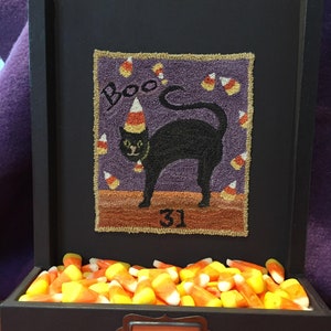 Puede incluir: Una caja de madera negra con un gato negro que lleva un gorro de fiesta y la palabra "Boo" sobre un fondo morado. La caja tiene un compartimento lleno de maíz dulce y una etiqueta que dice "Treats".