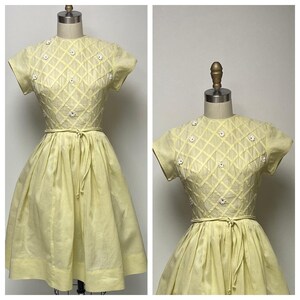 May include: A yellow vintage dress with short sleeves and a fitted bodice. The bodice is decorated with white embroidered flowers and a criss-cross pattern. The skirt is full and gathered at the waist. The dress has a tie at the waist.