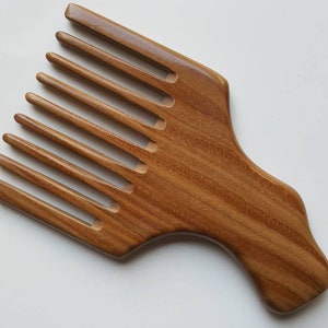 A wooden pick comb with a wide handle and a row of nicely spaced teeth. The comb is made of light brown wood with a natural grain pattern.