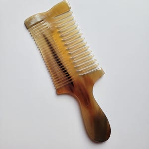 K&ouml;nnte beinhalten: Ein Haarkamm aus nat&uuml;rlichem Horn mit einem durchscheinenden, marmorierten Muster in Bernstein-, Creme- und Dunkelbraunt&ouml;nen. Der Kamm verf&uuml;gt &uuml;ber einen stabilen Griff und eine Reihe fein, gleichm&auml;&szlig;ig angeordneter Zinken.