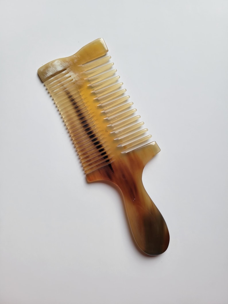 K&ouml;nnte beinhalten: Ein Haarkamm aus nat&uuml;rlichem Horn mit einem durchscheinenden, marmorierten Muster in Bernstein-, Creme- und Dunkelbraunt&ouml;nen. Der Kamm verf&uuml;gt &uuml;ber einen stabilen Griff und eine Reihe fein, gleichm&auml;&szlig;ig angeordneter Zinken.