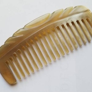 May include: A light brown comb made of horn with a carved leaf design. The comb has a rounded shape and is approximately 13 cm long.