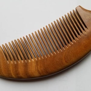 May include: A wooden comb with a curved handle and closely spaced teeth. The comb is made of a light brown wood and has a smooth, polished finish. A small hole is drilled near the top of the handle.