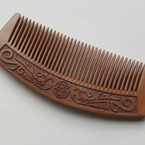 May include: A brown wooden comb with a carved design on the handle. The design features a swirl pattern and a circle with a symbol inside.